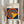 May include: Person in beige sweater holding framed Bauhaus poster with concentric circles in red, yellow, blue, and light blue on dark blue background, yellow triangle overlay. Light wood frame, cream matting. Dried pampas grass visible to left against white curtained window background.
