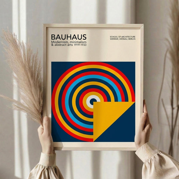 May include: Person in beige sweater holding framed Bauhaus poster with concentric circles in red, yellow, blue, and light blue on dark blue background, yellow triangle overlay. Light wood frame, cream matting. Dried pampas grass visible to left against white curtained window background.