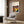 May include: Framed Bauhaus poster with colorful concentric circles in red, yellow, blue, and orange on navy background, displayed above a beige upholstered armchair with geometric patterned pillow. Modern living room setting features brass floor lamp, dark round side table with open book, wood paneled wall, and light wooden flooring.