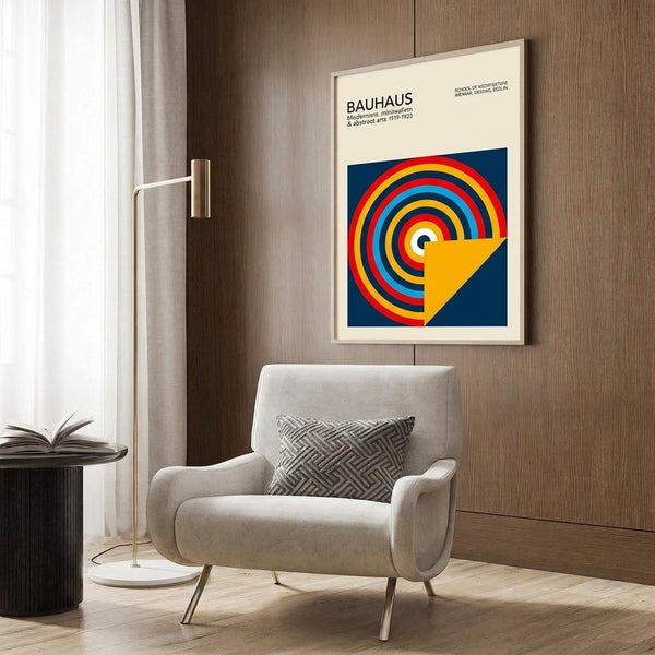 May include: Framed Bauhaus poster with colorful concentric circles in red, yellow, blue, and orange on navy background, displayed above a beige upholstered armchair with geometric patterned pillow. Modern living room setting features brass floor lamp, dark round side table with open book, wood paneled wall, and light wooden flooring.