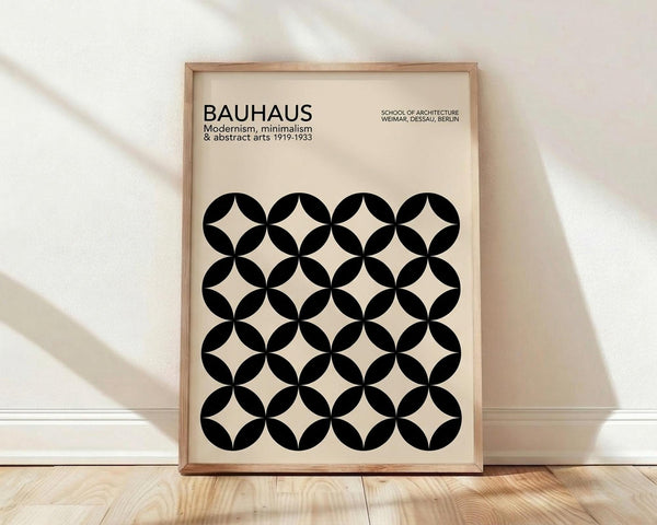 May include: Framed poster with cream background featuring black geometric pattern of overlapping circles creating diamond shapes between them. Text reads "BAUHAUS Modernism, minimalism & abstract arts 1919-1933" and "SCHOOL OF ARCHITECTURE WEIMAR, DESSAU, BERLIN" in black typography. Light wood frame leans against white wall on light wooden floor with soft natural lighting creating diagonal shadows.