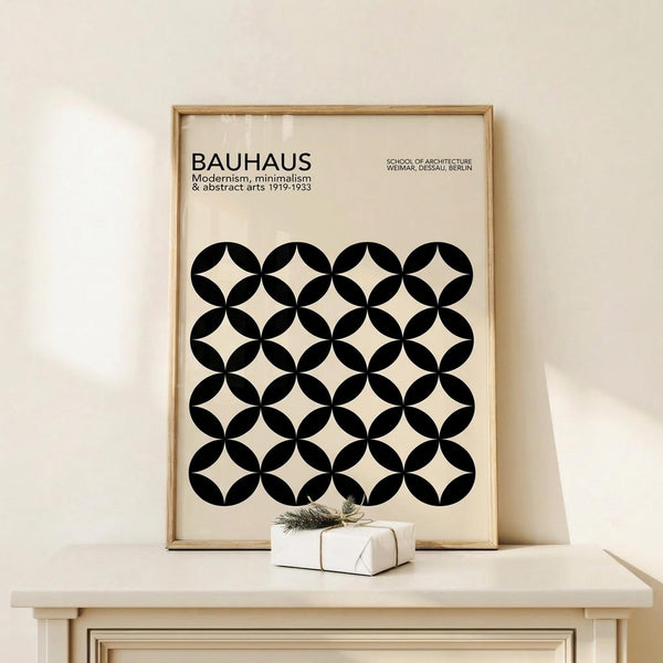 May include: Framed Bauhaus poster with black and white geometric circle pattern displayed on light wood shelf against cream wall. Modern minimalist art print in natural wood frame, accompanied by small wrapped gift with twine and greenery sprig.