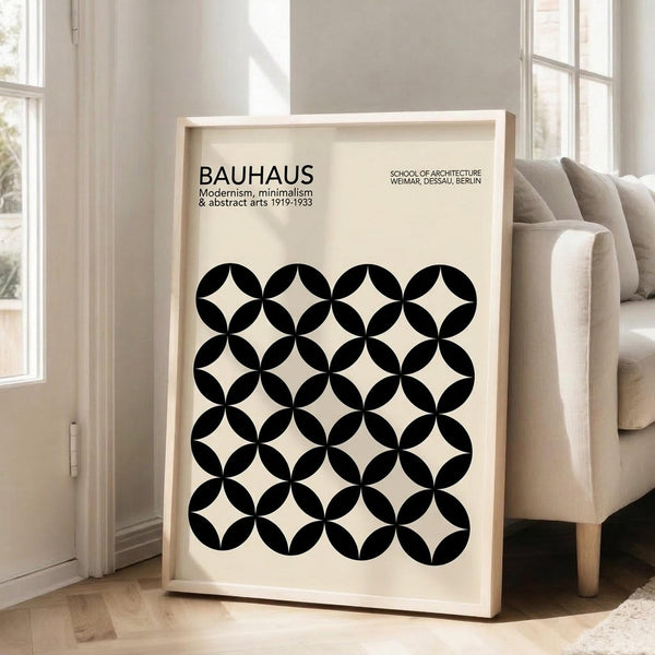 May include: Framed Bauhaus poster with geometric black circles pattern creating four-pointed star shapes on cream background, displayed in light wood frame leaning against white wall next to beige upholstered chair in bright living room with large windows and natural light streaming across hardwood floors.