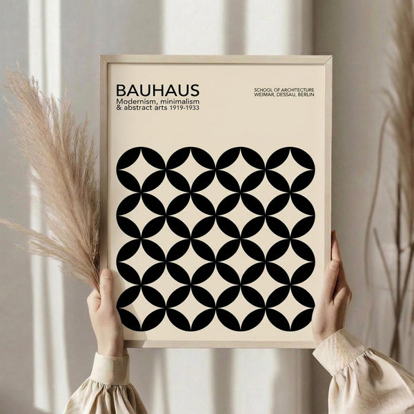 May include: Hands holding a framed Bauhaus-inspired poster with black circular overlapping pattern on cream background, displayed against white curtains with dried pampas grass visible to the left. The geometric design features intersecting circles creating diamond shapes between them. Light wood frame complements the minimalist aesthetic.
