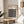 May include: Framed Bauhaus poster with geometric black circle pattern on cream background, displayed on round wooden side table next to beige upholstered chair. Natural lighting creates shadows across white wall. Brown paper bag and plant leaves visible in minimalist room setting.