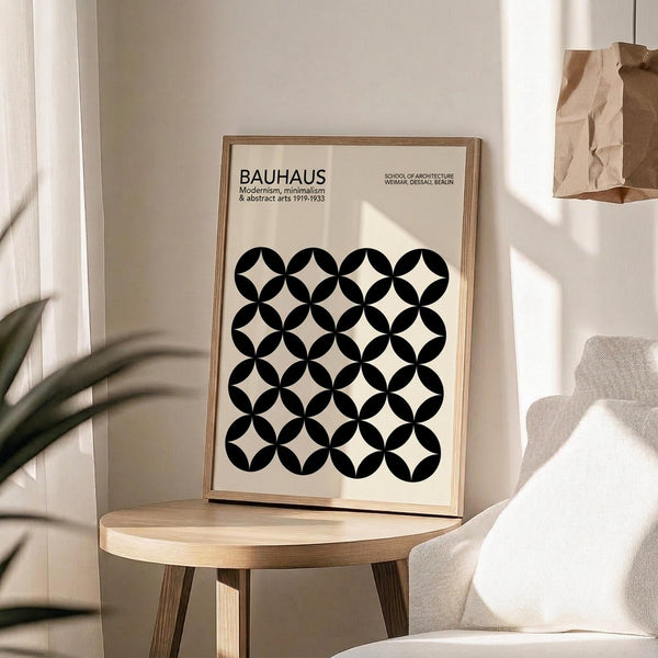 May include: Framed Bauhaus poster with geometric black circle pattern on cream background, displayed on round wooden side table next to beige upholstered chair. Natural lighting creates shadows across white wall. Brown paper bag and plant leaves visible in minimalist room setting.