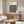 May include: Modern living room with cream walls featuring a large framed poster displaying black and white overlapping circles creating star-shaped patterns. Below sits a walnut wood credenza with sliding doors, white upholstered chair, glass-top oval coffee table, and copper accent lamp. Minimalist Scandinavian-style interior with natural lighting.
