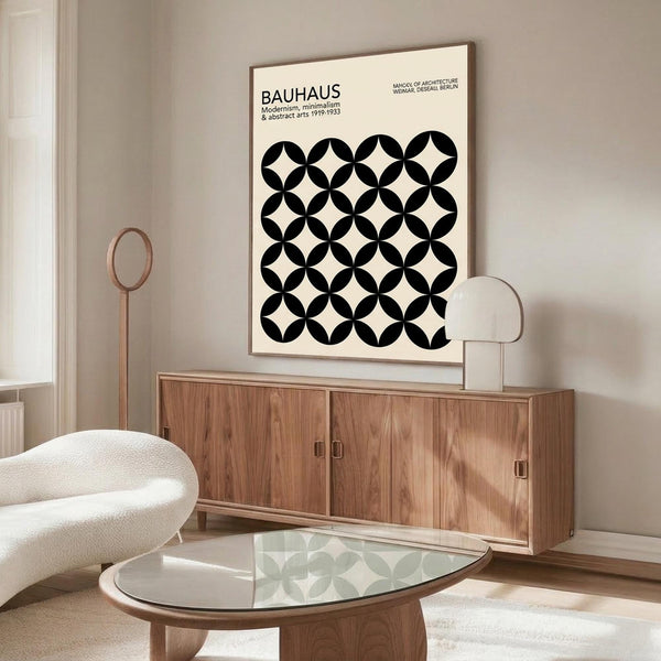 May include: Modern living room with cream walls featuring a large framed poster displaying black and white overlapping circles creating star-shaped patterns. Below sits a walnut wood credenza with sliding doors, white upholstered chair, glass-top oval coffee table, and copper accent lamp. Minimalist Scandinavian-style interior with natural lighting.