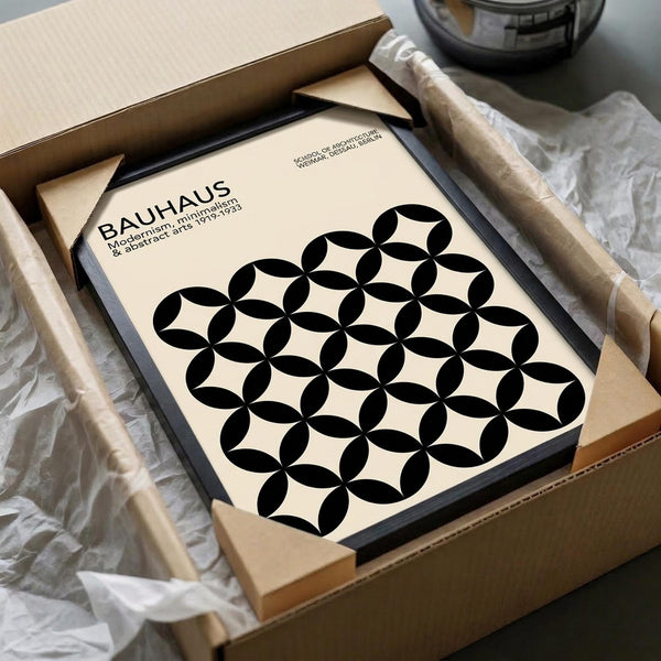 May include: Black framed poster with cream background featuring geometric pattern of overlapping black circles creating flower-like motifs, displayed in cardboard shipping box with protective wrapping materials. The minimalist design shows "BAUHAUS" text and dates "1919-1933" in upper left corner. Frame appears medium-sized relative to packaging materials visible around edges.