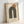 Minimalist geometric poster featuring olive green concentric arches with an orange-brown circle positioned above, printed on cream textured paper. Text reads "BAUHAUS 1919-1933 SCHOOL OF ARTS & CRAFTS" in brown lettering. The artwork is displayed in a light wood frame with hanging hardware, leaning against a white wall on a light surface.