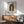 May include: Mid-century modern living room with sage green armchair and wooden sideboard. Framed Bauhaus poster featuring geometric design with dark green concentric arcs, terracotta circle, and cream background. Natural sunlight creates shadows across white walls. Round wooden coffee table and ceramic table lamp complete the minimalist scene.