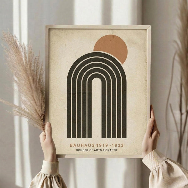 May include: Person holding framed Bauhaus-inspired poster with geometric design featuring dark green concentric arches and terracotta circle on cream textured background, displayed next to dried pampas grass in neutral toned room setting