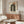 May include: Bauhaus-inspired poster with black concentric arches and orange circle on cream background, displayed above wooden sideboard in modern living room. White curved chair and glass coffee table visible. Poster features clean geometric design with mid-century aesthetic. Room has neutral tones with natural wood furniture and minimalist white table lamp.
