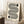 May include: Framed Bauhaus-style poster with black geometric lines forming flowing, striped patterns and circular elements on cream background, displayed against white wall next to wooden side table with coffee cup and beige textile, creating minimalist home decor scene with natural lighting and shadows