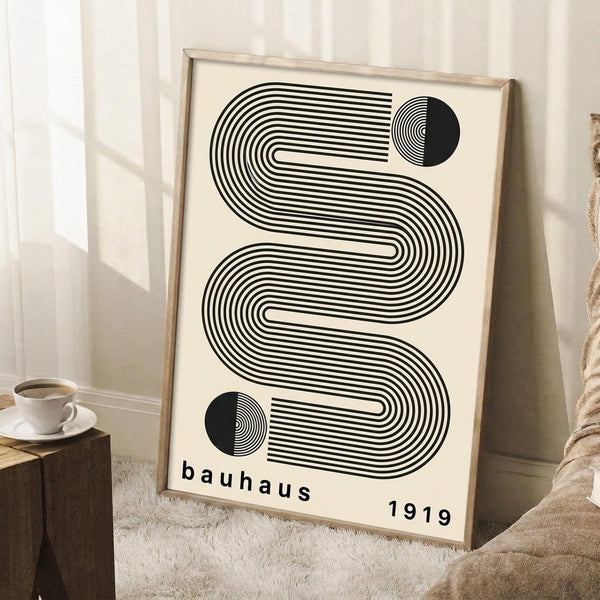 May include: Framed Bauhaus-style poster with black geometric lines forming flowing, striped patterns and circular elements on cream background, displayed against white wall next to wooden side table with coffee cup and beige textile, creating minimalist home decor scene with natural lighting and shadows