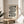 May include: Black and white geometric poster with curved linear design and two black circles, featuring "bauhaus 1919" text at bottom, displayed in thin wooden frame on round wooden side table next to beige upholstered chair with plant leaves visible in soft natural lighting