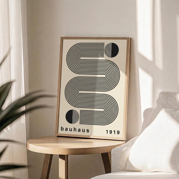 May include: Black and white geometric poster with curved linear design and two black circles, featuring "bauhaus 1919" text at bottom, displayed in thin wooden frame on round wooden side table next to beige upholstered chair with plant leaves visible in soft natural lighting