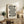 May include: Black and white geometric poster with flowing curved lines and circular elements displaying "bauhaus 1919" text, shown in light wood frame leaning against white wall on rustic wooden sideboard. Room features neutral beige chair, fern plant, ceramic vases, and books creating warm minimalist interior styling.