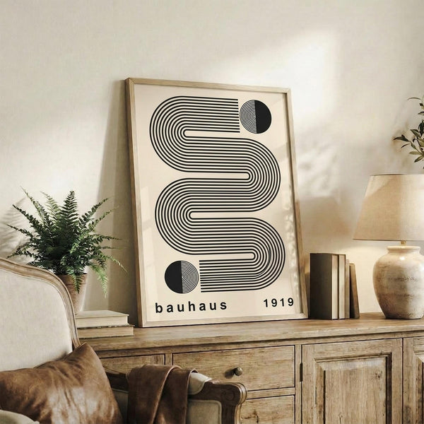 May include: Black and white geometric poster with flowing curved lines and circular elements displaying "bauhaus 1919" text, shown in light wood frame leaning against white wall on rustic wooden sideboard. Room features neutral beige chair, fern plant, ceramic vases, and books creating warm minimalist interior styling.