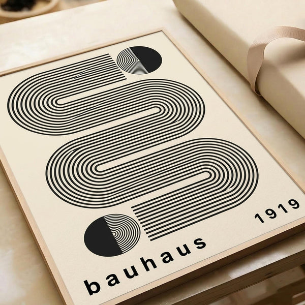 May include: Framed Bauhaus-style poster with black concentric oval lines forming geometric pattern on cream background, featuring two half-circles with radiating lines, displayed on neutral beige surface with rolled paper tube nearby. Modern minimalist design with "bauhaus 1919" text at bottom.