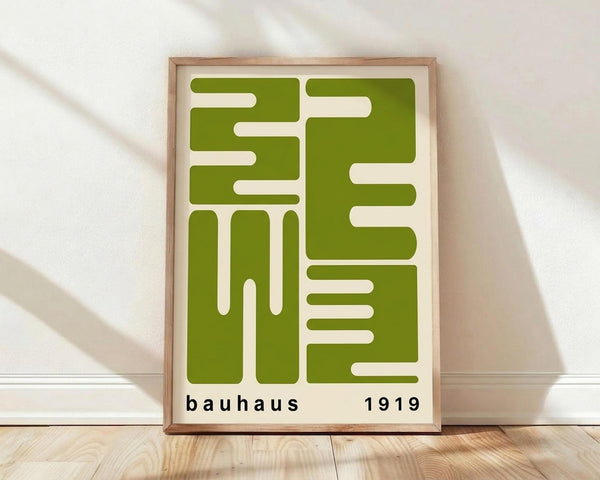 May include: Framed geometric poster with olive green abstract shapes on cream background, displayed against white wall on light wood floor. Modern minimalist design features interlocking rectangular and linear forms. Text reads "bauhaus 1919" in black lowercase letters at bottom. Natural wood frame with visible grain pattern. Soft shadows cast on textured white fabric backdrop.