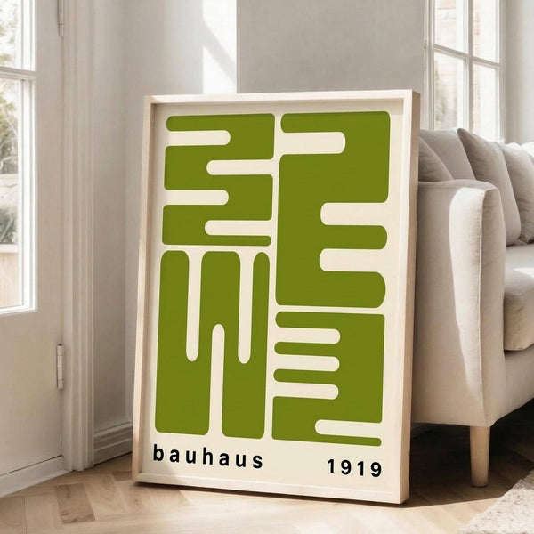 May include: Green geometric Bauhaus-style poster in light wood frame leaning against white wall in bright living room. Chunky beige sectional sofa with throw pillows visible to right. White window frames with natural light streaming in. Hardwood floors with area rug. Poster features bold lime green angular shapes on cream background with "bauhaus 1919" text at bottom.