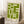 May include: Framed green and cream geometric Bauhaus-style poster leaning against white wall in bright room. Modern abstract design features interlocking rectangular shapes with "bauhaus 1919" text at bottom. Natural wood frame, positioned on fluffy white rug next to wooden side table with coffee cup and saucer. Soft natural lighting creates shadows on wall.