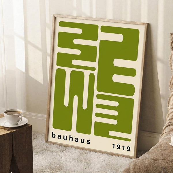 May include: Framed green and cream geometric Bauhaus-style poster leaning against white wall in bright room. Modern abstract design features interlocking rectangular shapes with "bauhaus 1919" text at bottom. Natural wood frame, positioned on fluffy white rug next to wooden side table with coffee cup and saucer. Soft natural lighting creates shadows on wall.