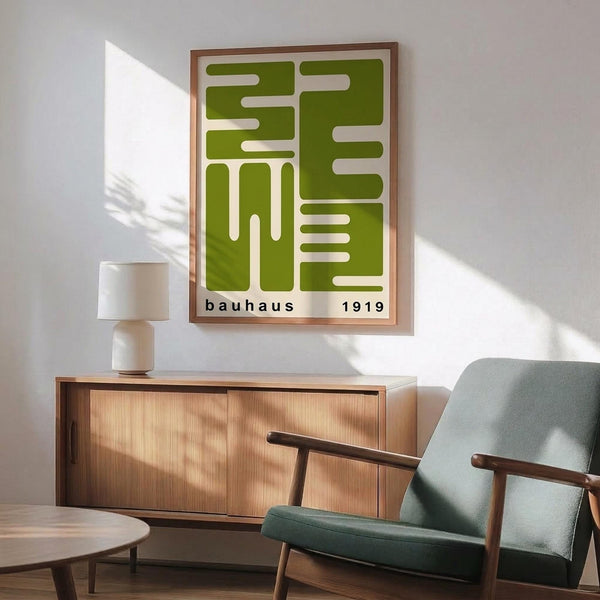 May include: Green geometric Bauhaus poster with white abstract shapes and "bauhaus 1919" text in wooden frame, displayed above wood credenza with white lamp. Mid-century modern living room setting features sage green upholstered armchair with wooden arms, round coffee table, white walls with natural sunlight shadows.