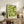 May include: Green geometric Bauhaus poster with "1919" text displayed in light wood frame on rustic wooden sideboard. Scene shows cream armchair, fern plant, ceramic vase, stacked books, and neutral wall background in cozy living room setting.