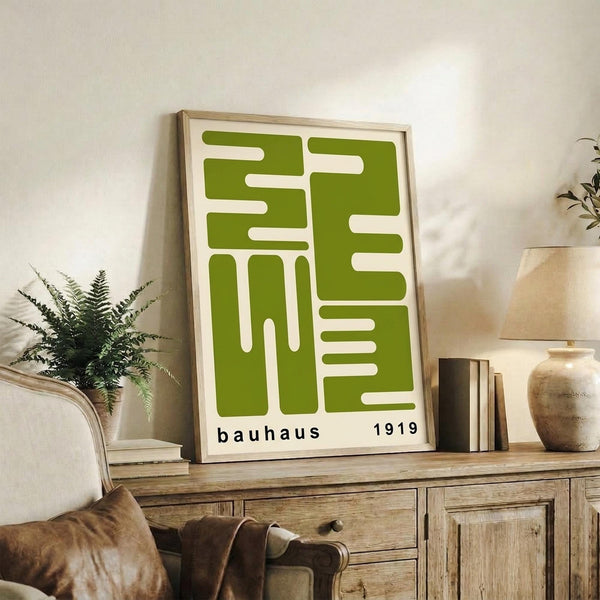 May include: Green geometric Bauhaus poster with "1919" text displayed in light wood frame on rustic wooden sideboard. Scene shows cream armchair, fern plant, ceramic vase, stacked books, and neutral wall background in cozy living room setting.