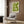 May include: Green geometric poster with white linear pattern in light wood frame, displayed on wood-paneled wall above light gray upholstered armchair with geometric throw pillow, alongside brass floor lamp and dark round side table with open book