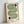 May include: Framed geometric art print leaning against white wall. Features flowing green striped wave pattern with two black half-circles containing concentric lines. Text reads "bauhaus 1919" in lowercase black letters on cream background. Light wood frame casts soft shadow on wall.