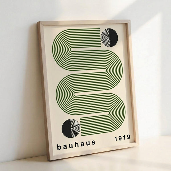 May include: Framed geometric art print leaning against white wall. Features flowing green striped wave pattern with two black half-circles containing concentric lines. Text reads "bauhaus 1919" in lowercase black letters on cream background. Light wood frame casts soft shadow on wall.