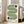 May include: Framed poster with green wavy lines and black half-circles on cream background, displayed in bright living room corner next to beige armchair and large windows with natural light streaming in