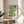 May include: Framed geometric poster with green flowing wave design and black circular elements on cream background, displayed on round wooden stool next to beige upholstered chair, with plant leaves visible in soft natural lighting against neutral wall