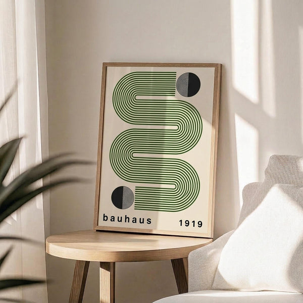 May include: Framed geometric poster with green flowing wave design and black circular elements on cream background, displayed on round wooden stool next to beige upholstered chair, with plant leaves visible in soft natural lighting against neutral wall