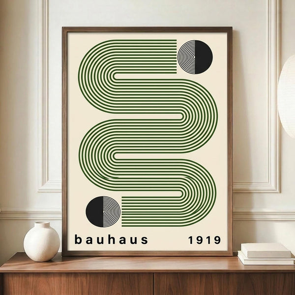 May include: Framed geometric poster with green flowing wavy lines and two black semicircles, displayed on wooden sideboard next to white ceramic vase and books. The artwork features parallel curved stripes creating an S-shaped pattern on cream background, with "bauhaus 1919" text at bottom. Modern mid-century interior setting with white paneled walls and warm wood furniture.