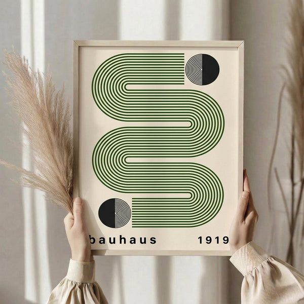 May include: Person in cream sweater holding framed geometric art print featuring flowing green and black striped wave pattern with circular accents, displayed against neutral curtained background with dried pampas grass bundle, showcasing minimalist Bauhaus-inspired design in natural wood frame