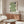 May include: Modern living room with wooden sideboard displaying framed geometric poster featuring flowing green striped wave design with black circular elements on cream background. Room styled with contemporary white curved chair, glass coffee table, and minimalist decor in neutral tones.