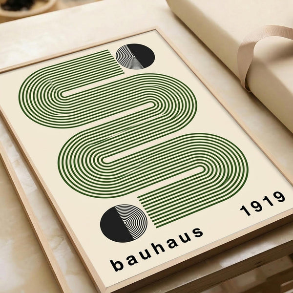 May include: Framed poster with geometric design featuring flowing green concentric lines forming wave-like patterns against cream background, two black circular elements with radiating lines, "bauhaus 1919" text at bottom. Wooden frame sits on light wooden surface next to rolled beige paper tube, natural lighting creates soft shadows.