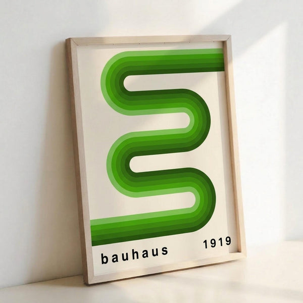 May include: Framed poster with green gradient serpentine design creating flowing S-shaped curves against cream background, displayed in light wood frame leaning against white wall. Black text reads "bauhaus 1919" at bottom. Modern minimalist Bauhaus-inspired geometric art print shown in home setting with soft natural lighting creating gentle shadows.
