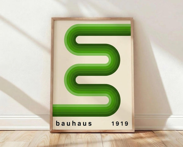 May include: Framed poster with green wavy serpentine design on cream background, displaying "bauhaus 1919" text below. Wood frame leaning against white wall on light wooden floor with soft natural lighting and shadows.