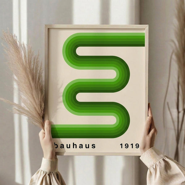 May include: Person in beige sweater holding framed poster with green curved geometric ribbons forming flowing S-shape design. Black text reads "bauhaus 1919" below the vibrant green layered bands. Light wooden frame against cream background with dried pampas grass visible on left side.