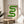 May include: Framed poster with green wavy geometric design in Bauhaus style, displayed on round wooden side table next to beige upholstered chair. Natural lighting creates soft shadows on neutral wall. Green ribbonlike form creates flowing S-curve against cream background with "bauhaus 1919" text below.