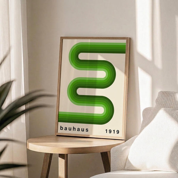 May include: Framed poster with green wavy geometric design in Bauhaus style, displayed on round wooden side table next to beige upholstered chair. Natural lighting creates soft shadows on neutral wall. Green ribbonlike form creates flowing S-curve against cream background with "bauhaus 1919" text below.