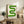 May include: Green geometric poster with curved S-shaped design in gradual color transition from light to dark green, displayed in light wood frame on rustic wooden sideboard. Room setting shows neutral beige chair, potted fern, ceramic vases, and books creating warm modern interior styling against cream wall.