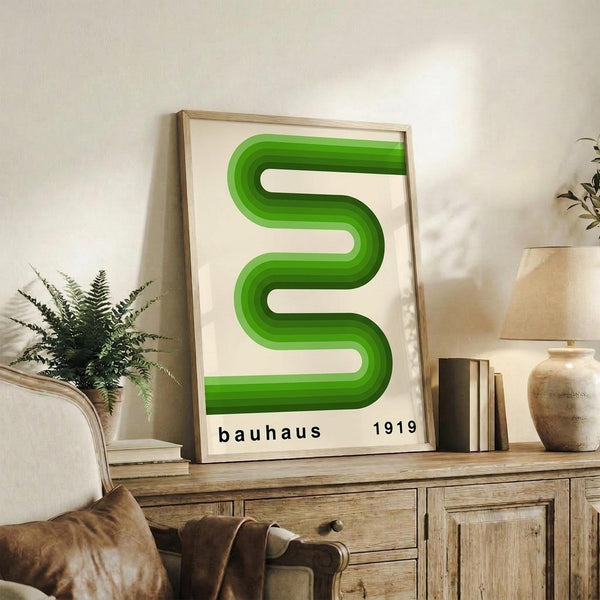 May include: Green geometric poster with curved S-shaped design in gradual color transition from light to dark green, displayed in light wood frame on rustic wooden sideboard. Room setting shows neutral beige chair, potted fern, ceramic vases, and books creating warm modern interior styling against cream wall.
