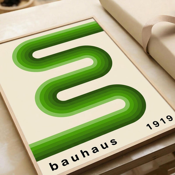 May include: Green modernist poster with flowing S-shaped curves in gradient shades from light to dark green, displayed in light wood frame on cream colored surface. Text reads "bauhaus" and "1919" in black lowercase letters. Minimalist geometric design with layered dimensional effect. Rolled white paper visible in background.