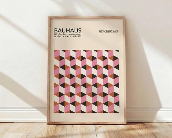 May include: Framed Bauhaus poster with geometric cube pattern in pink, orange, black and cream colors, displayed in wooden frame against white wall on light wood floor with natural window lighting creating soft shadows